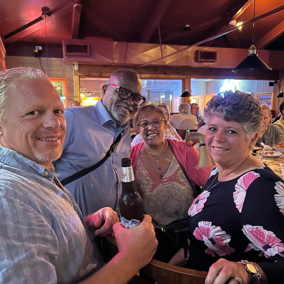 Four smiling friends pose together inside a warmly lit restaurant, with tables, diners, pendant lights, and a beer bottle visible.