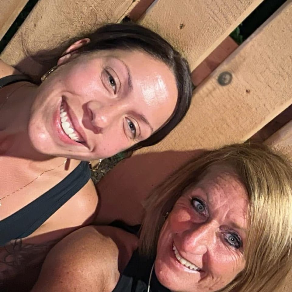 Two smiling women pose for a selfie outdoors against a wooden fence.