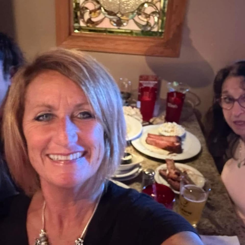 Four women smile for a selfie at a restaurant table with plates of food, drinks, and festive decor.