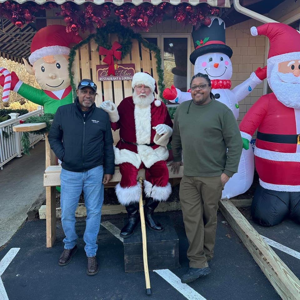 Two men pose beside Santa and inflatable Christmas decorations outside a restaurant adorned with festive garlands and a striped awning.