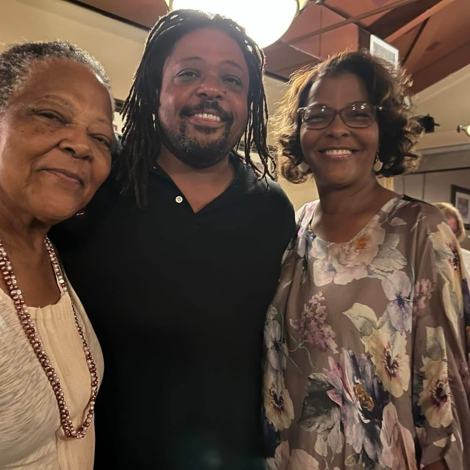 Three smiling adults pose together inside a warmly lit restaurant, with framed artwork and other diners in the background.