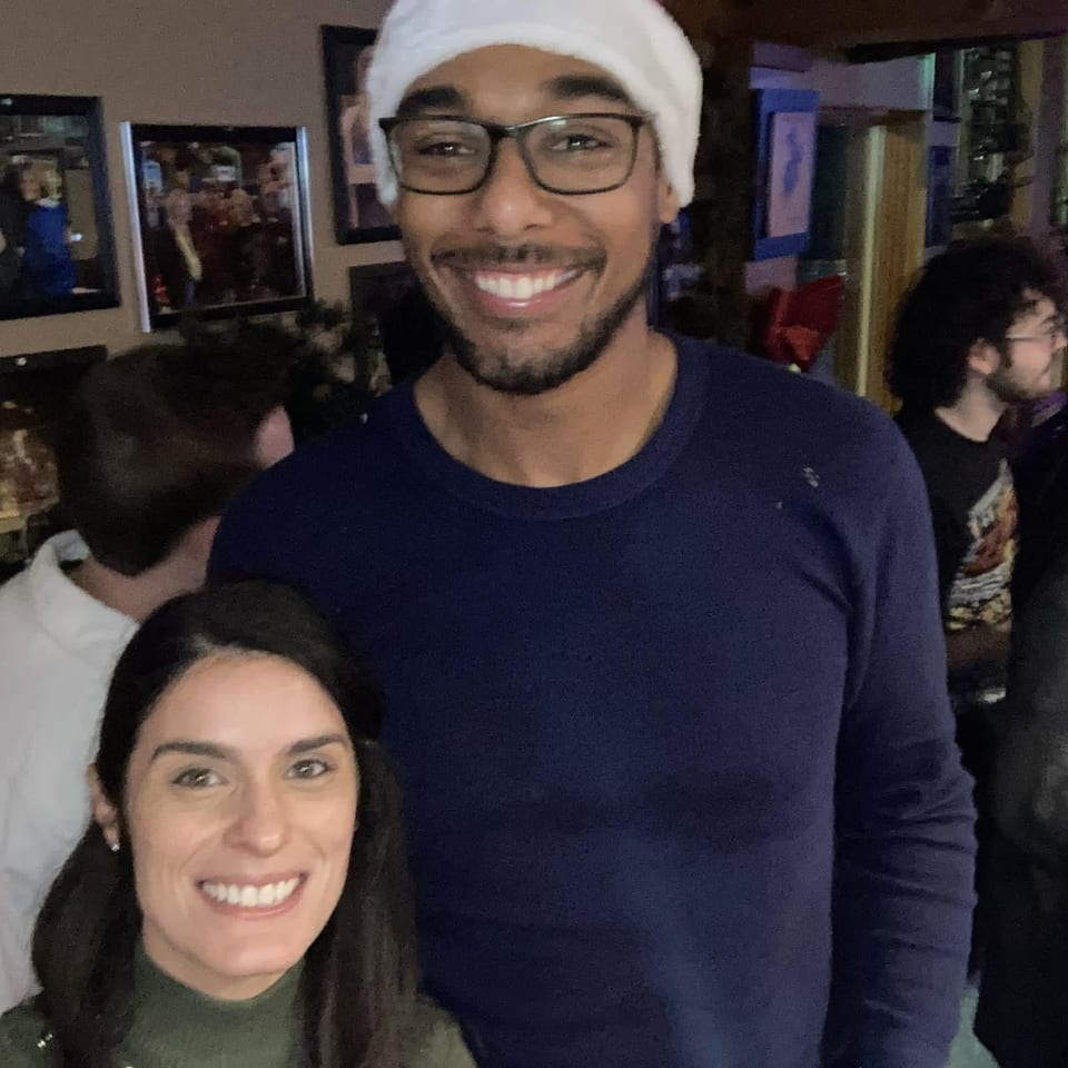 Smiling man in glasses and white hat poses with a woman at a lively, dimly lit restaurant gathering.