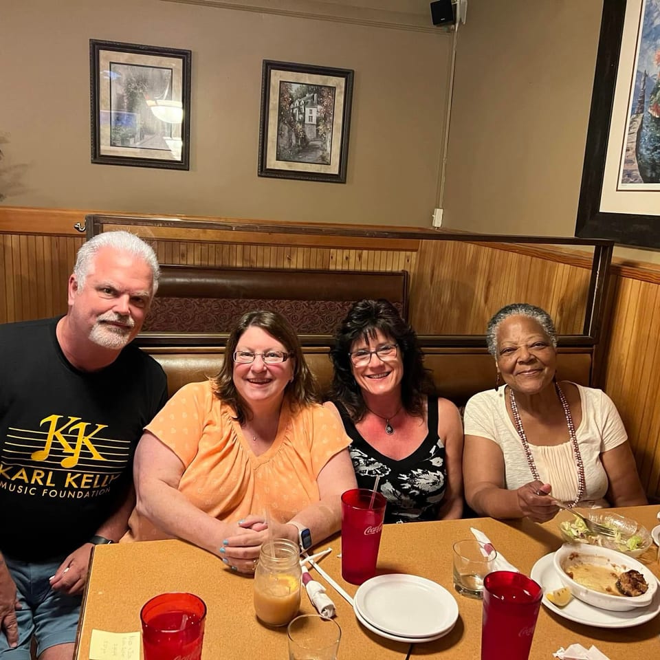Four people smile together in a restaurant booth surrounded by framed artwork, wood paneling, drinks, and partially eaten meals.