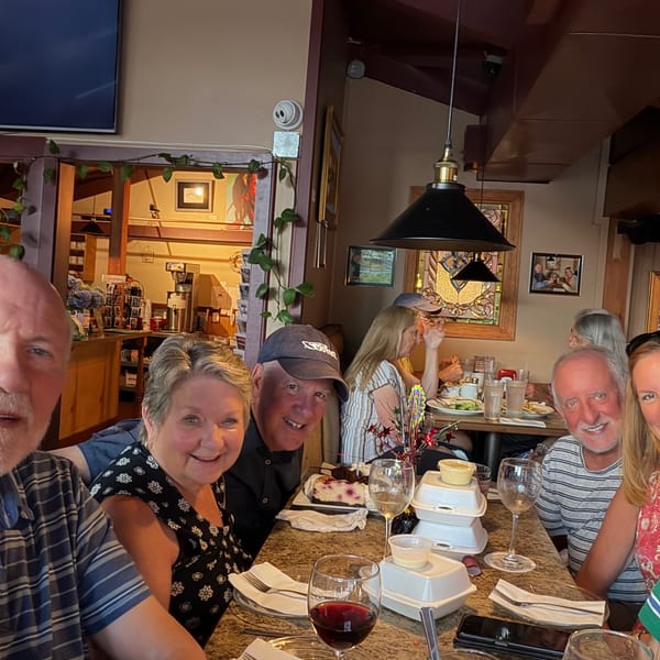 Six friends smile for a selfie around a restaurant table with drinks and meals in a warmly decorated dining room.