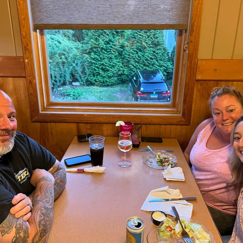 Four diners smile around a restaurant table with drinks and salads, seated beside a window overlooking a parked car and trees.