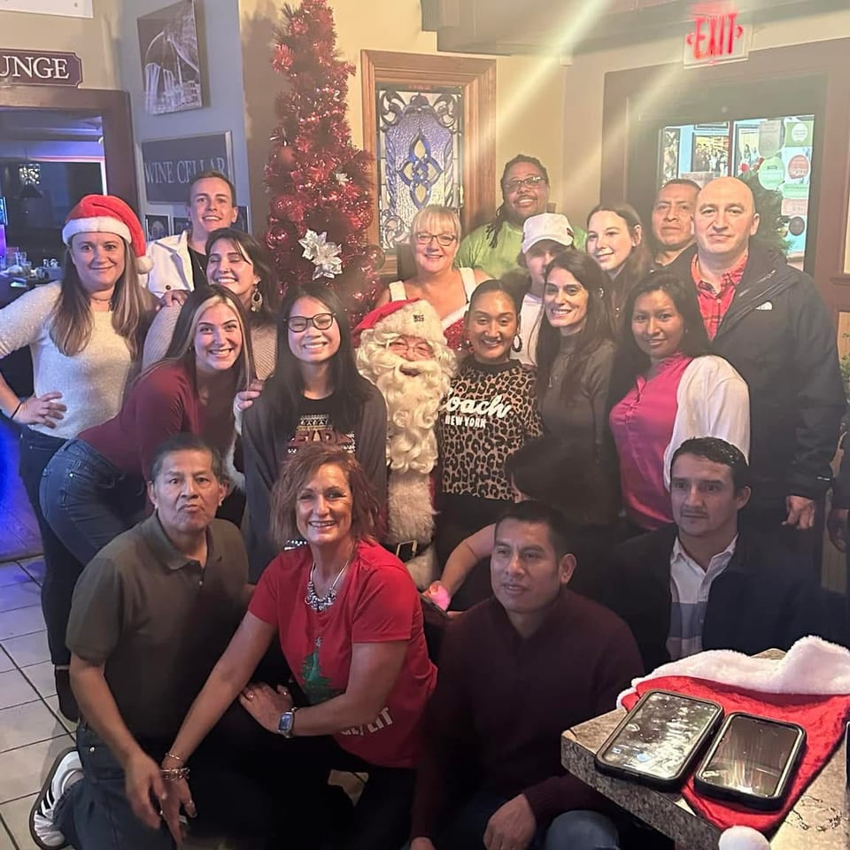 Large group posing indoors beside a decorated Christmas tree, with a person dressed as Santa and festive decorations.