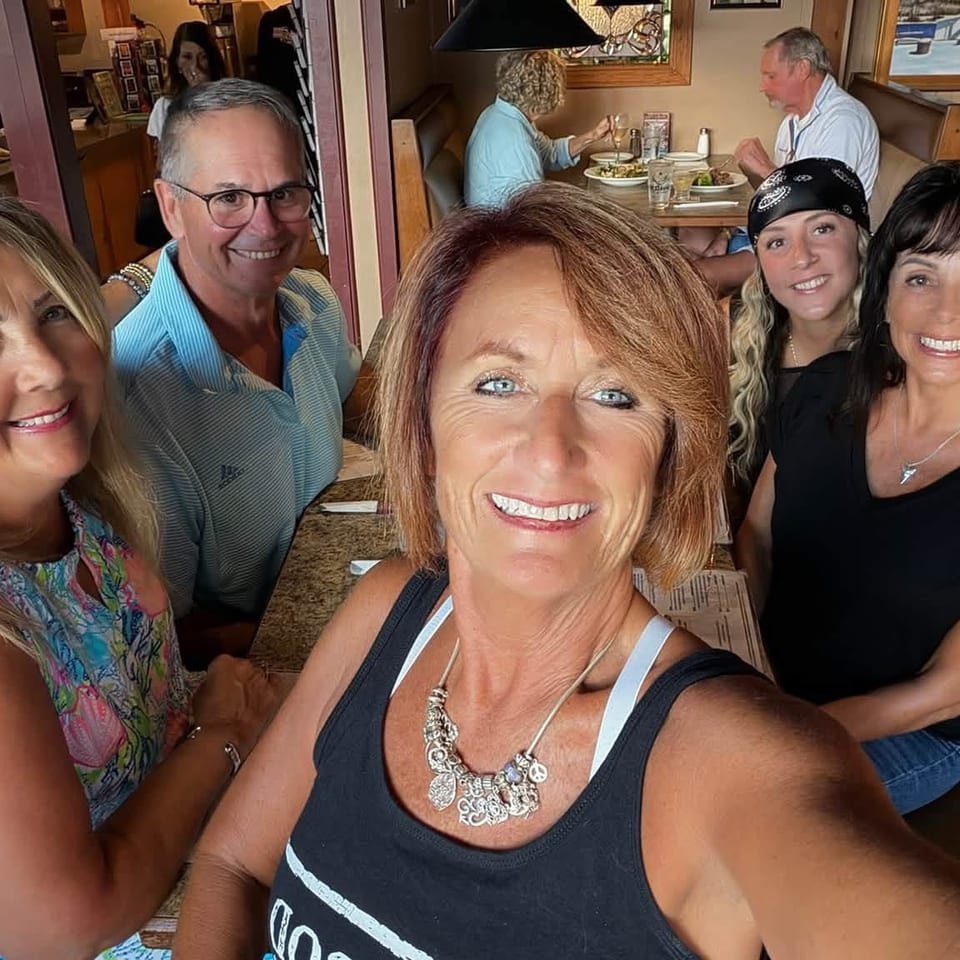 Five friends smile for a selfie inside a warmly decorated restaurant, with diners and meals visible in the background.