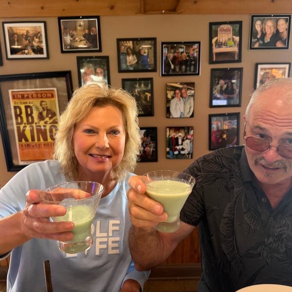 Two smiling diners raise pale green cocktails inside a restaurant decorated with framed photographs and memorabilia.