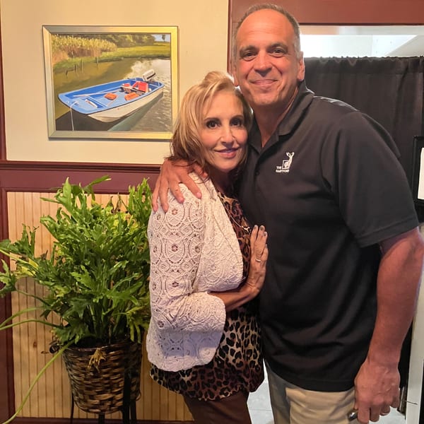 Smiling couple embrace inside a restaurant beside a potted plant and framed painting of a boat.