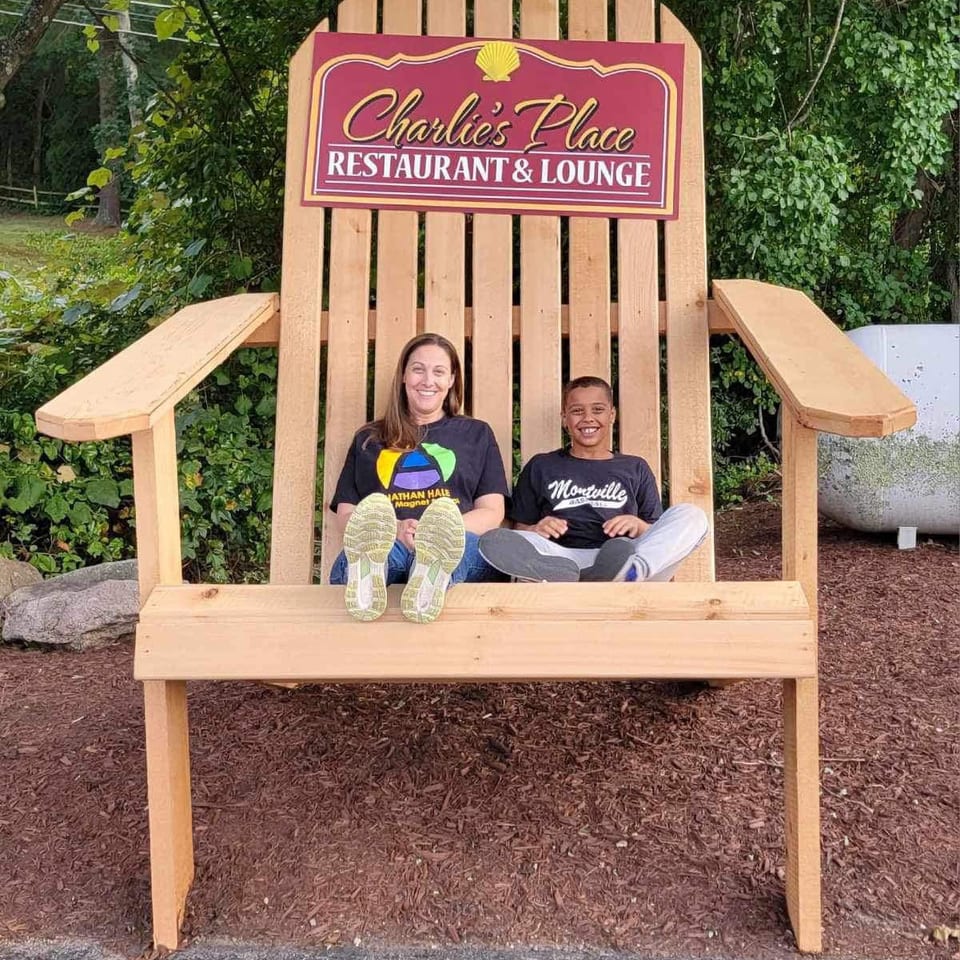 Two people sit on a giant Adirondack chair beneath a Charlie’s Place Restaurant & Lounge sign, surrounded by greenery.