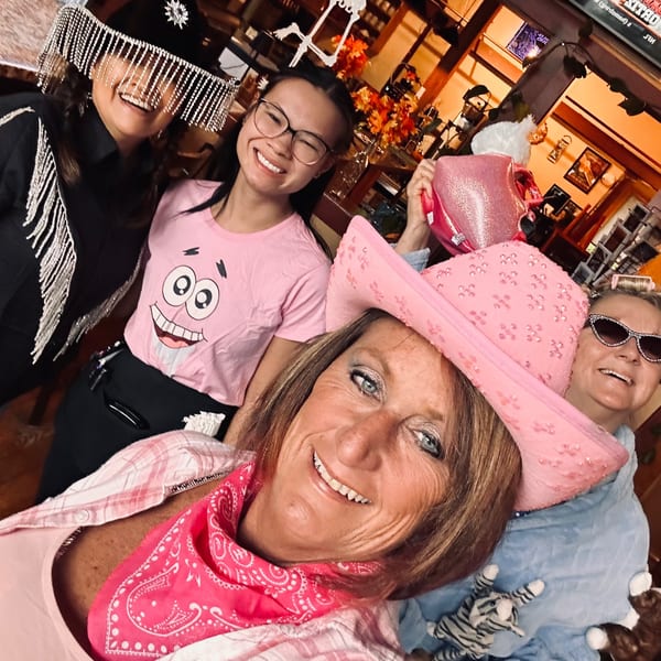 Friends pose for a cheerful selfie inside a warmly lit restaurant decorated with Halloween skeletons and costumes.