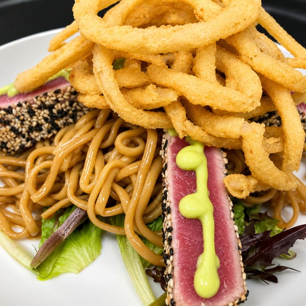 Sesame-crusted seared tuna with soy noodles, greens, and crispy fried onions on a white plate.