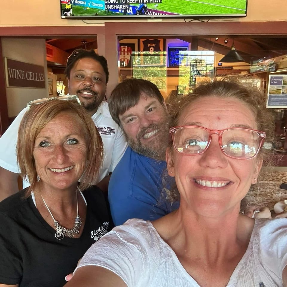 Four smiling people pose for a selfie inside a warmly decorated restaurant, with a wine cellar sign, televisions, and plated food visible.