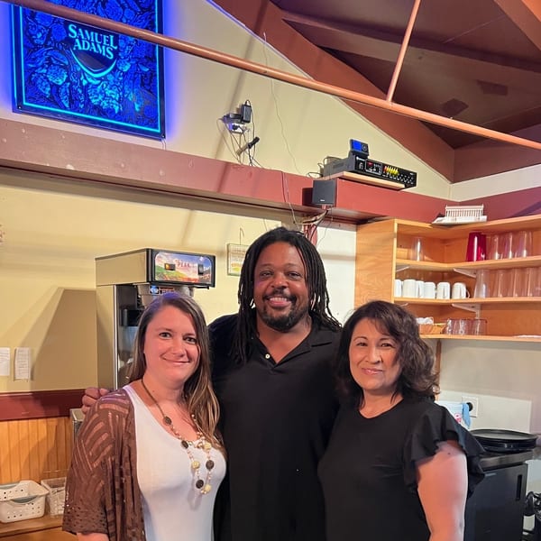 Three smiling people pose together inside a warmly lit restaurant near a coffee station and glassware shelves.