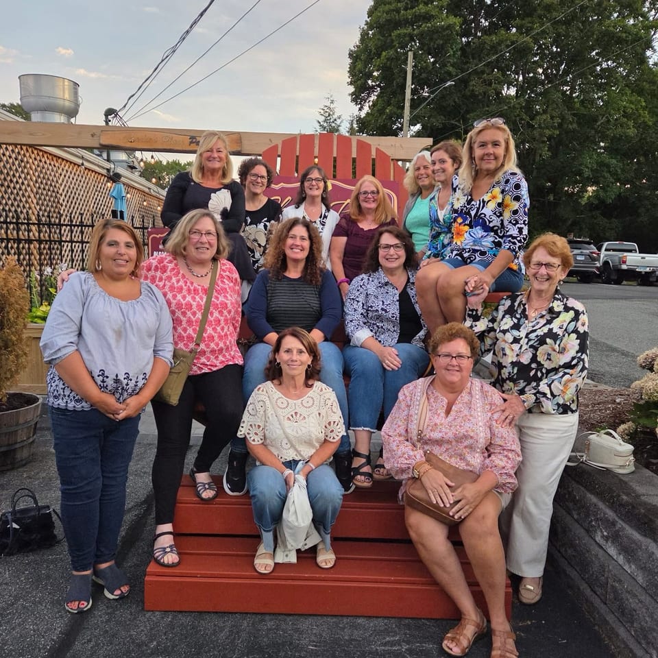 A large group of women poses on red outdoor benches beside a restaurant patio, with trees and utility wires behind them.