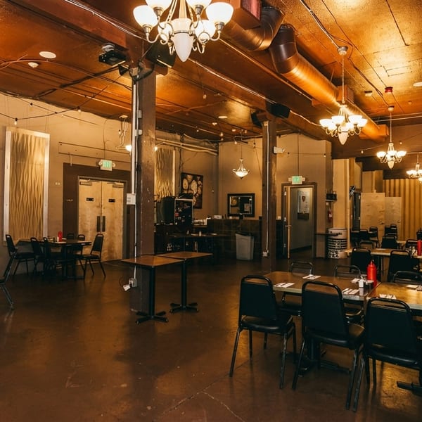Spacious restaurant dining room with dark tables and chairs, exposed brick, and chandeliers.