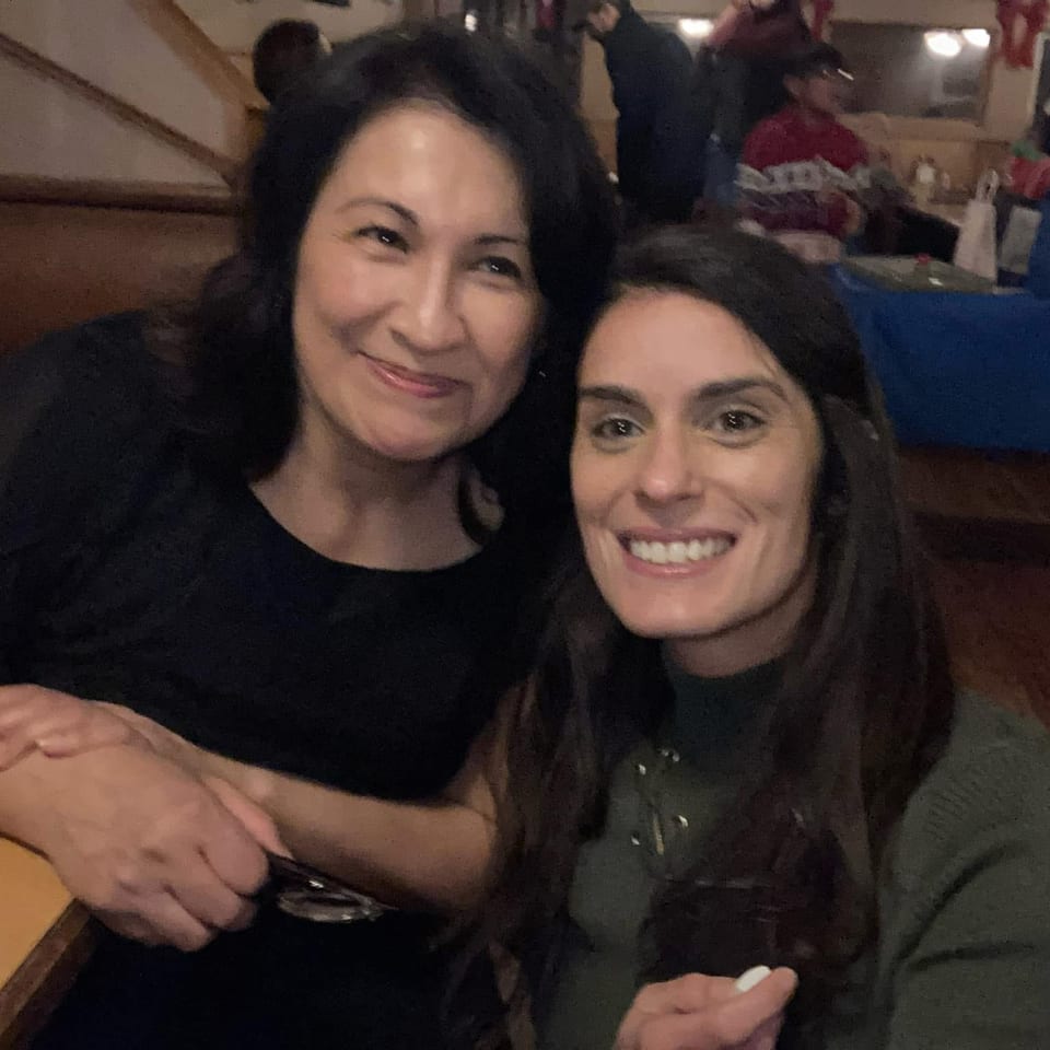 Two smiling women pose together in a cozy restaurant booth, with festive decorations and other diners in the background.