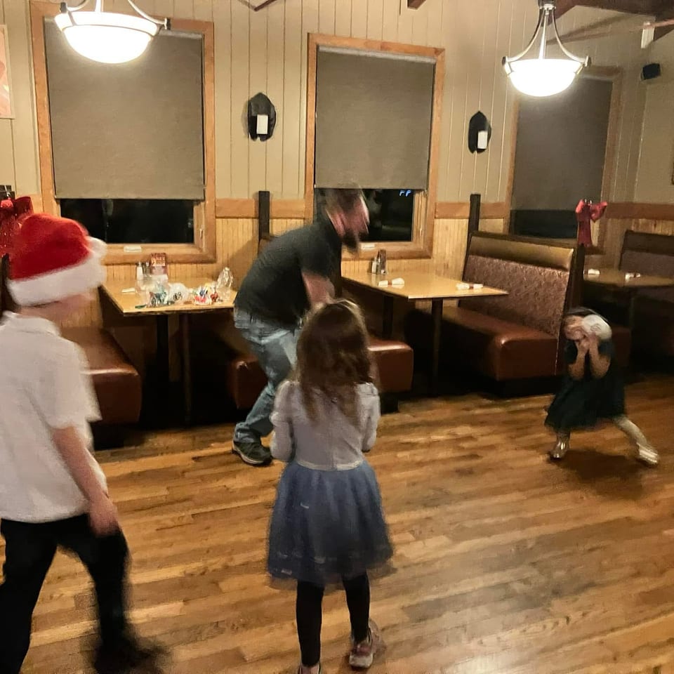 Children and an adult gather on a restaurant’s wooden floor, with one child wearing a Santa hat and another crouching near booths.