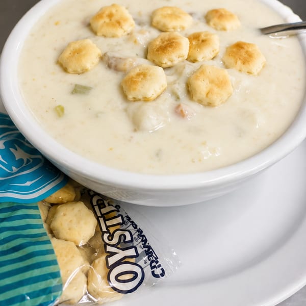Creamy clam chowder in a white bowl topped with oyster crackers, with a packet of crackers beside it.