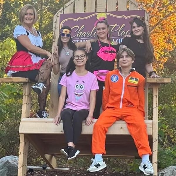 Five people in colorful costumes pose on a large wooden chair beneath a theatrical sign surrounded by autumn trees.