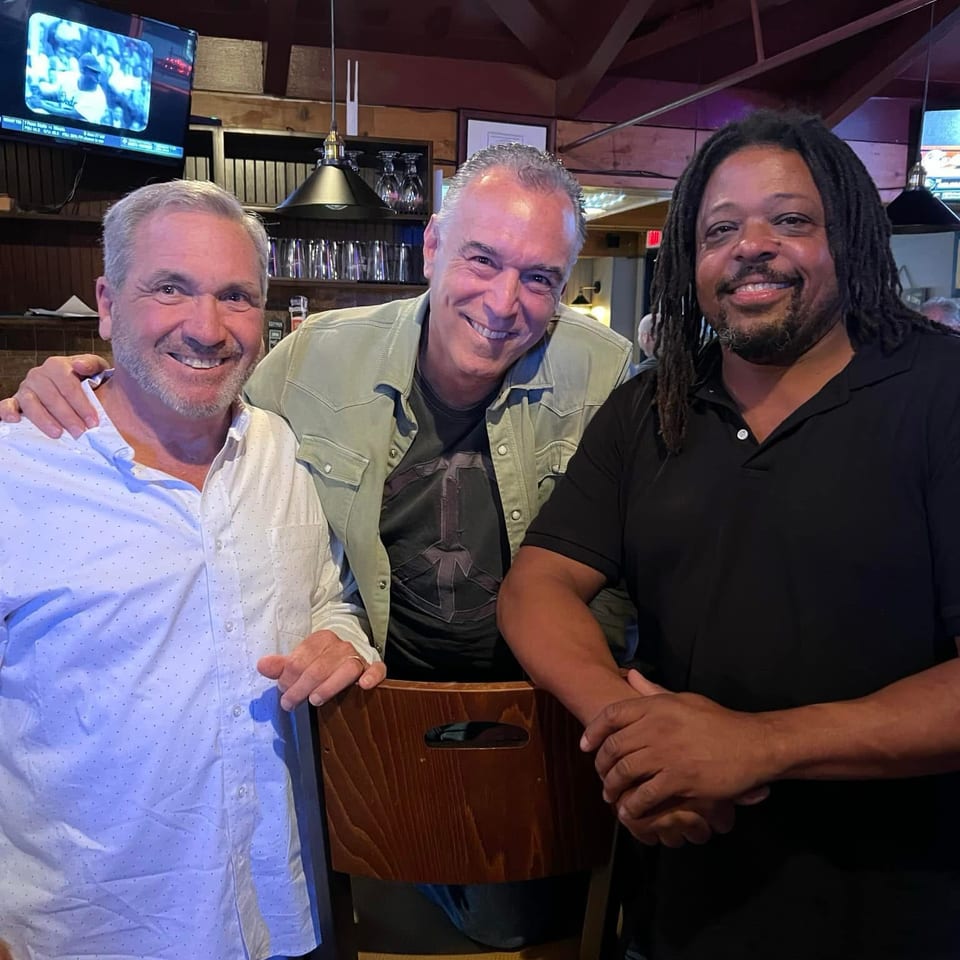 Three smiling men pose together inside a warmly lit restaurant bar, with pendant lights, glassware, and televisions visible behind them.