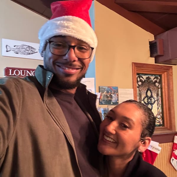 Smiling couple pose together inside a warmly decorated restaurant, with Christmas stockings, a decorated tree, and lounge signage behind them.