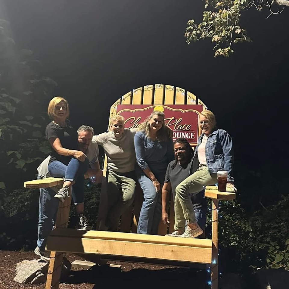 Six adults pose on a wooden bench beneath a colorful lounge sign, surrounded by trees and outdoor lighting.