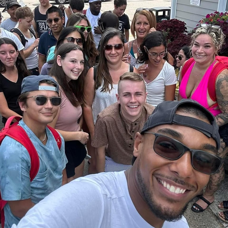 Large group smiling for a selfie beside outdoor restaurant tables and colorful flower planters.