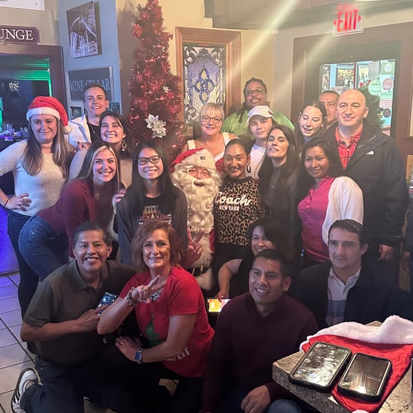 Large group poses together inside a restaurant beside a decorated Christmas tree, with several people wearing Santa hats.