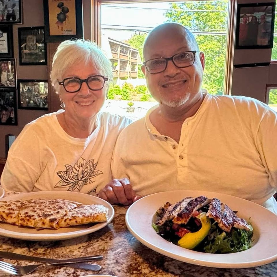 Smiling couple seated in a restaurant beside plates of flatbread and grilled meat salad, with framed photographs and sunny windows behind them.