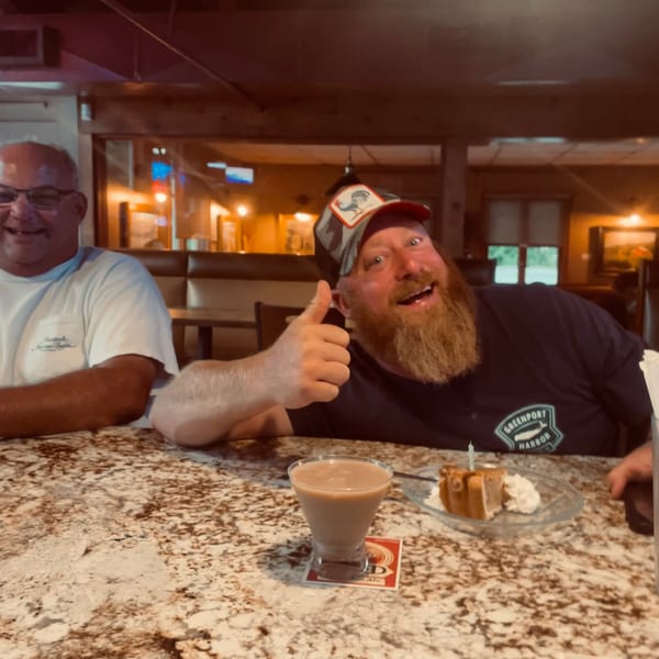Two men smile at a restaurant counter, one giving a thumbs-up beside a dessert plate and creamy drink.