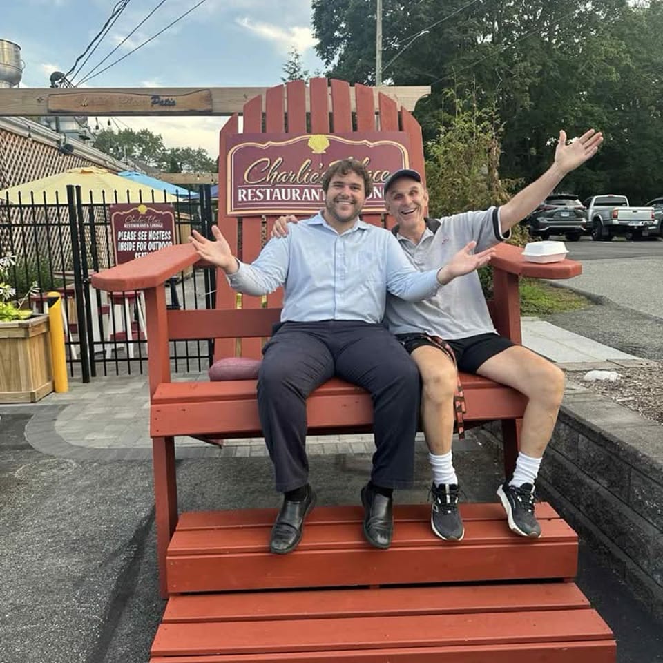 Two men pose on an oversized red bench outside Charlie’s Place restaurant, with the restaurant sign, patio, trees, and parking lot behind them.